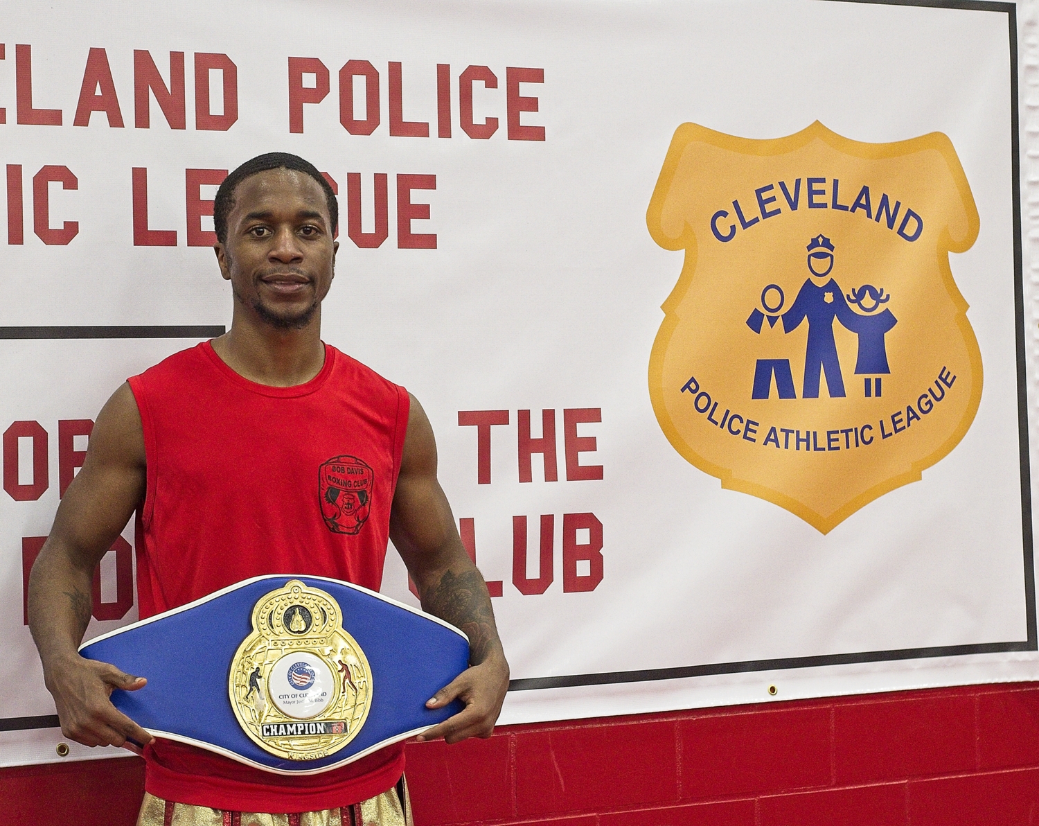 A young man displays his Champion belt while standing in front of the Cleveland Police Athletic League logo