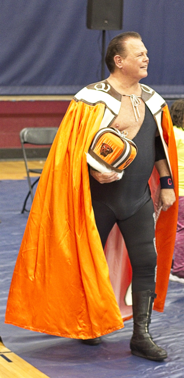 WWE Hall Of Fame Wrestler Jerry "The King" Lawler wearing a black wrestling singlet, sporting a Cleveland Guardians wristband and wearing a Cleveland Browns orange/brown/white cape. He carries an ornate orange/brown/white crown covered with Dawg emblems.